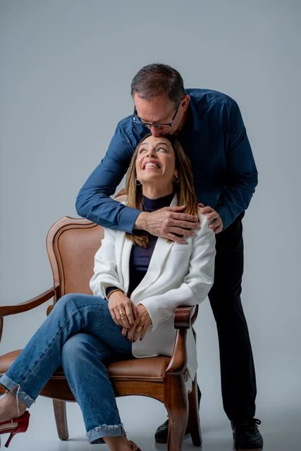 Dra. Mônica sentada na poltrona e Dr. Marcelo inclinado ao lado dela, sorrindo